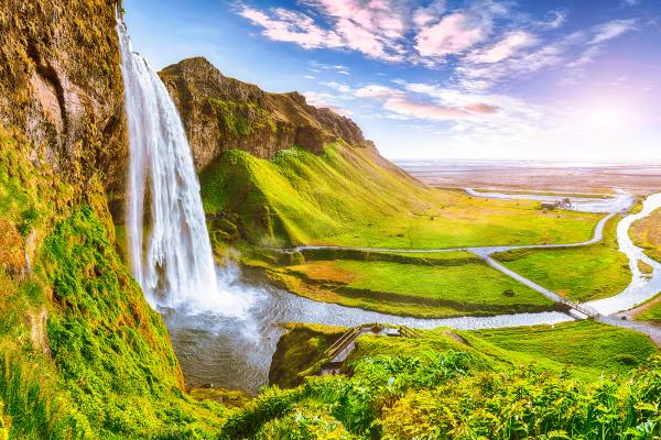 Seljalandsfoss waterfall plunging from a green cliff into a winding river in a lush valley under a vibrant sky.