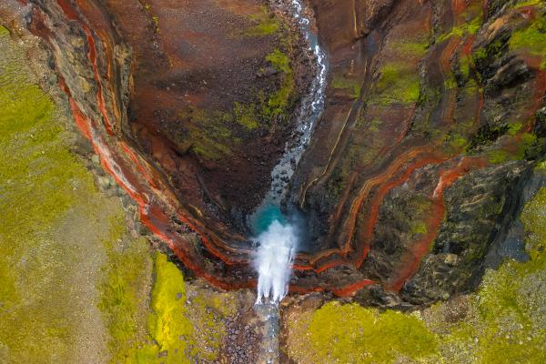 Aerial view of a vibrant canyon with a waterfall, turquoise pool, reddish rock layers, and green moss.
