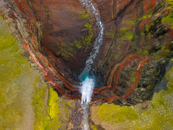 Overhead view of a narrow canyon with vibrant red, brown, and green layered walls, and a waterfall plunging into a turquoise pool.