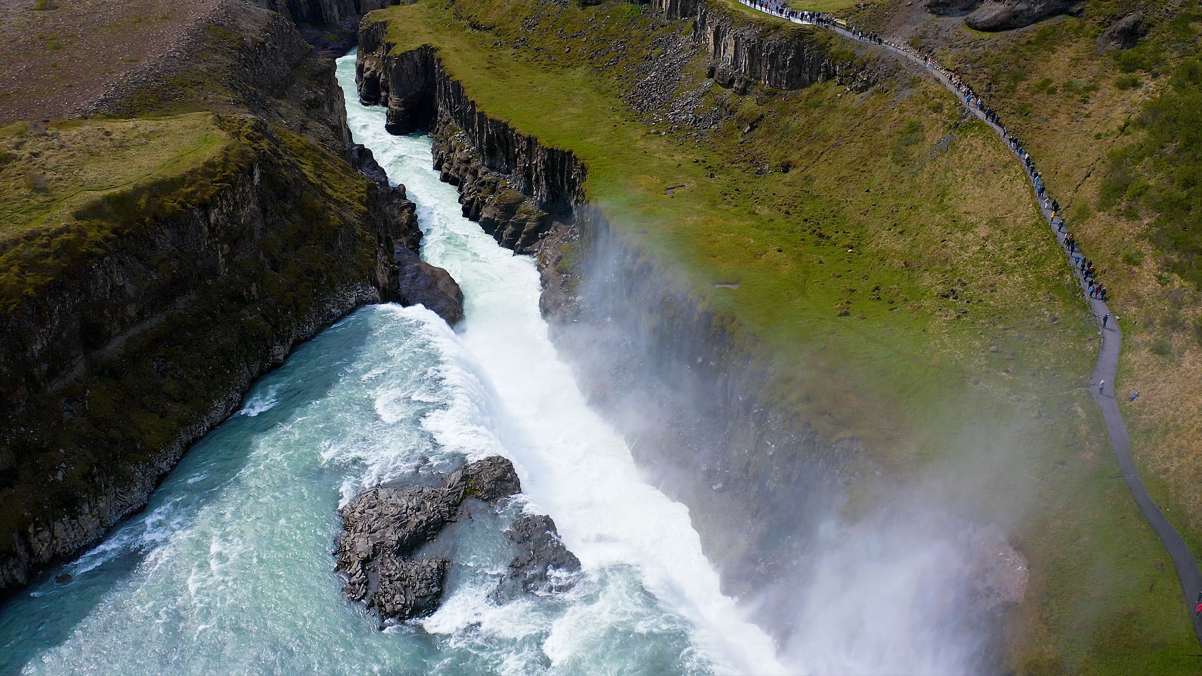 Aerial view of a powerful waterfall in a canyon, with a line of people on a path along the grassy cliff.