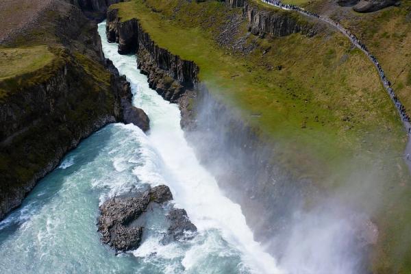 Aerial view of a powerful waterfall in a canyon, with a line of people on a path along the grassy cliff.