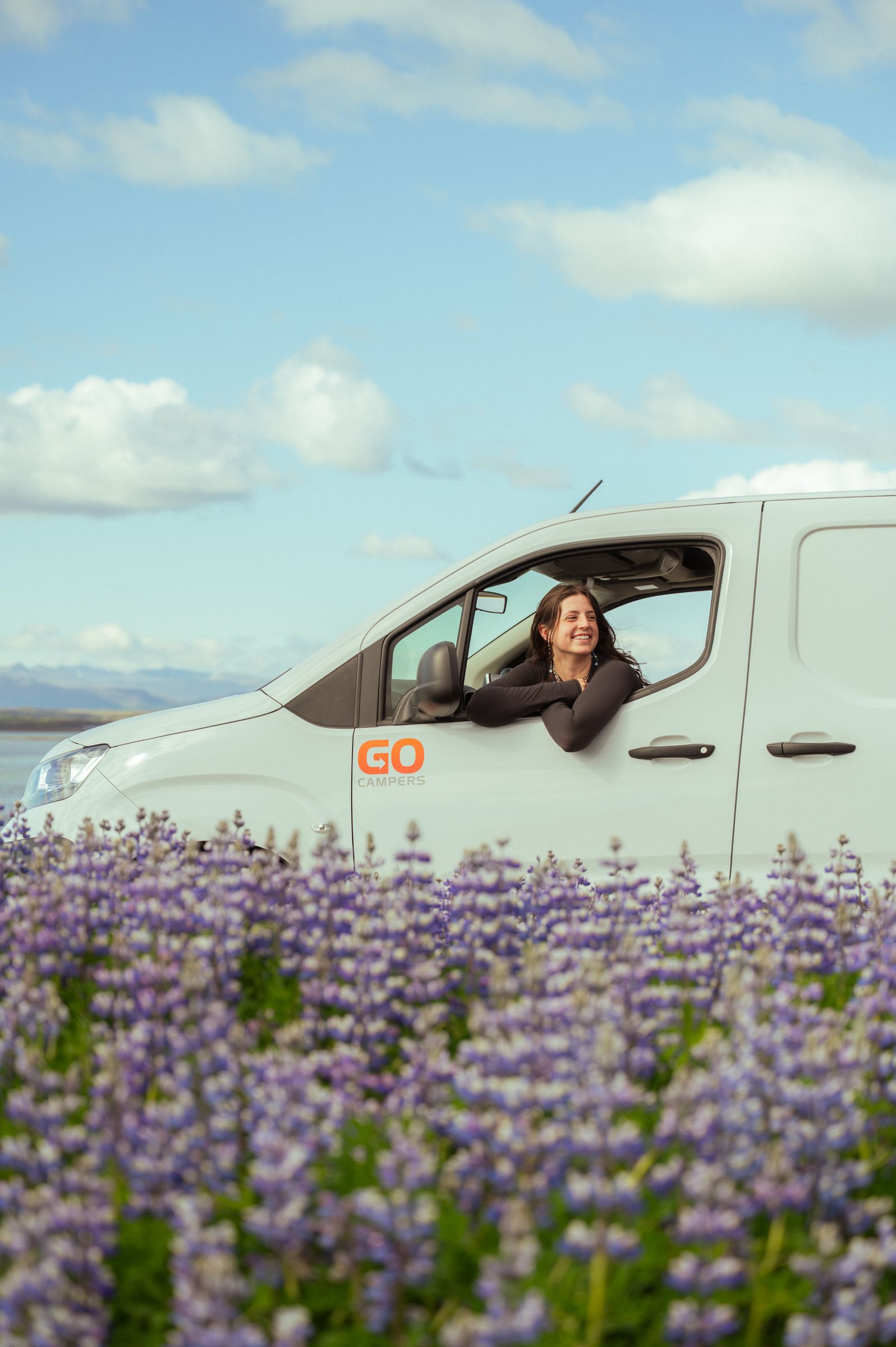 A smiling woman leans out of a white "GO CAMPERS" van parked in a field of purple flowers, with a lake and mountains in the distance under a blue sky.