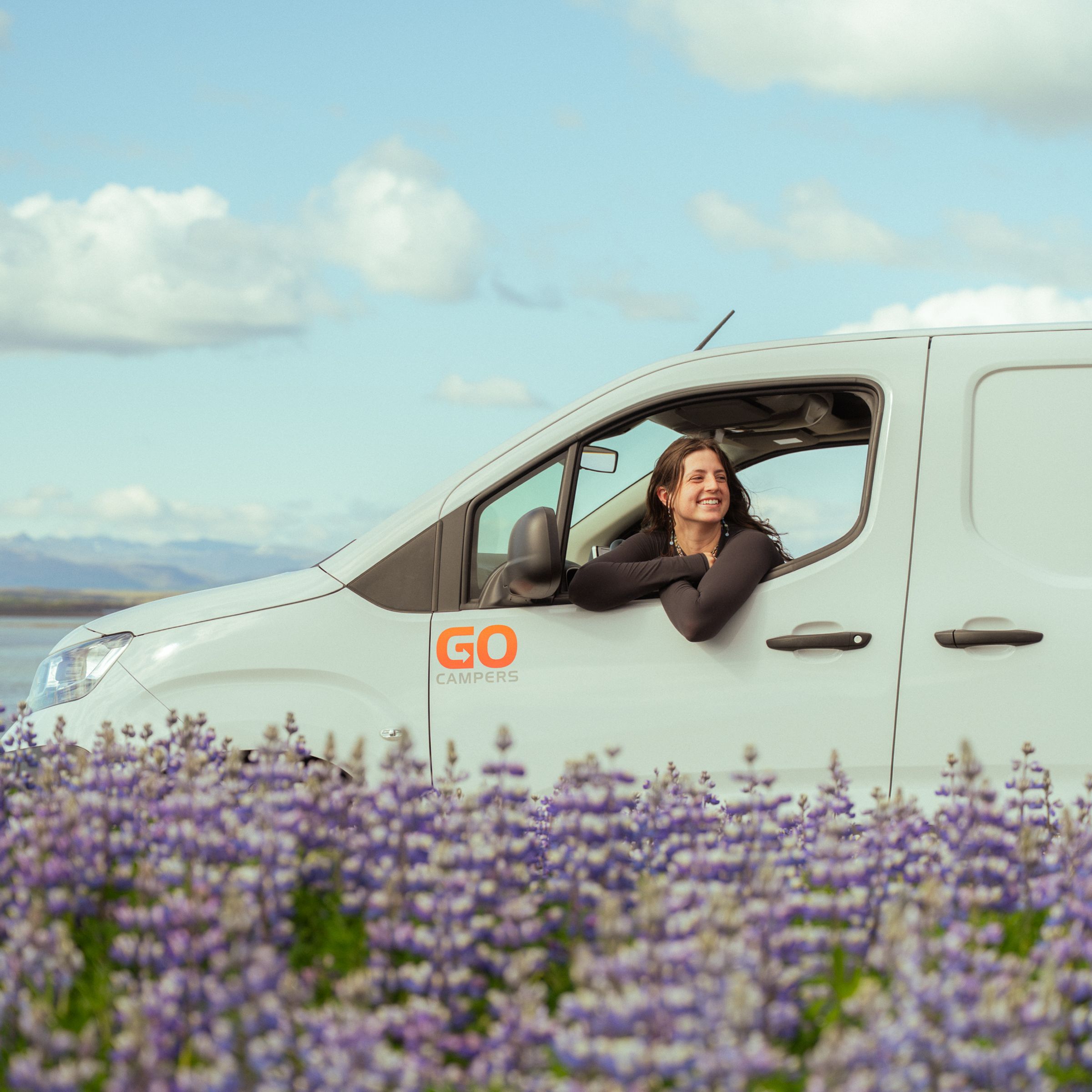 A smiling woman leans out of a white "GO CAMPERS" van parked in a field of purple flowers, with a lake and mountains in the distance under a blue sky.