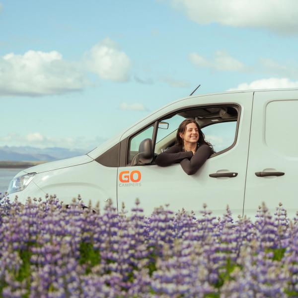 A smiling woman leans out of a white "GO CAMPERS" van parked in a field of purple flowers, with a lake and mountains in the distance under a blue sky.