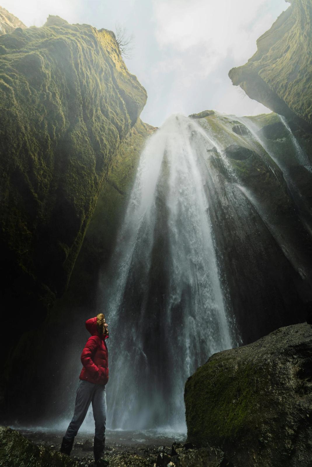 Person in a red jacket looking up at a powerful waterfall cascading down a mossy canyon.