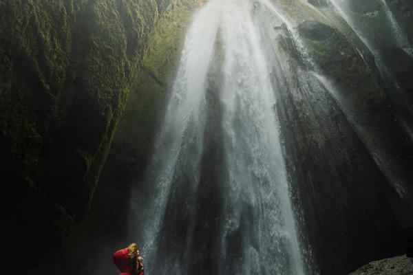 Person in a red jacket looking up at a powerful waterfall cascading down a mossy canyon.