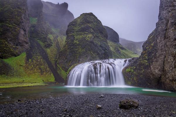 A smooth waterfall flows into a turquoise pool, flanked by dark, mossy cliffs and a gravel foreground.