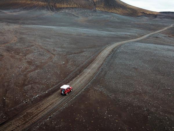 A red 4x4 drives on a dirt road through a dark, rocky volcanic landscape with hills.