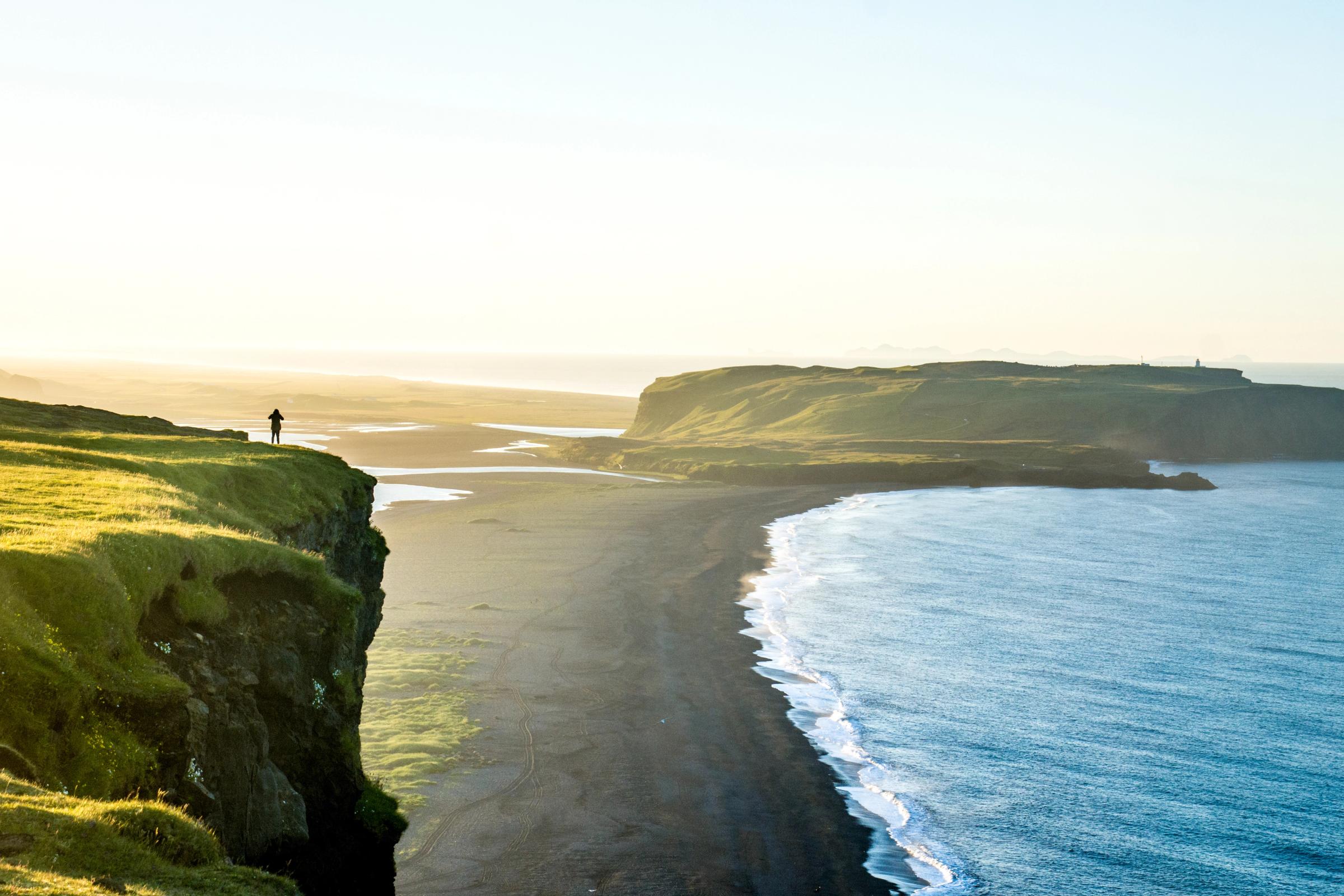 A solitary person stands on a grassy cliff, gazing out at a vast black-sand beach and an ocean bathed in the golden light of sunset.