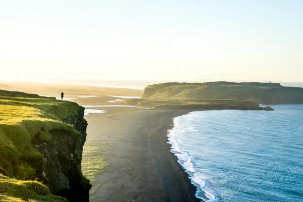 A solitary person stands on a grassy cliff, gazing out at a vast black-sand beach and an ocean bathed in the golden light of sunset.