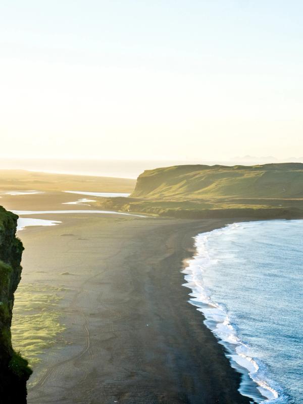 Una persona está de pie en un acantilado cubierto de hierba, contemplando una playa de arena negra y el océano bajo un cielo dorado.