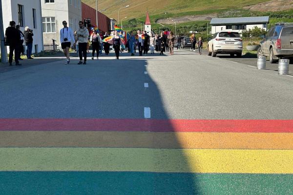 People march on a street with a prominent rainbow crosswalk in the foreground, surrounded by town buildings and green hills.