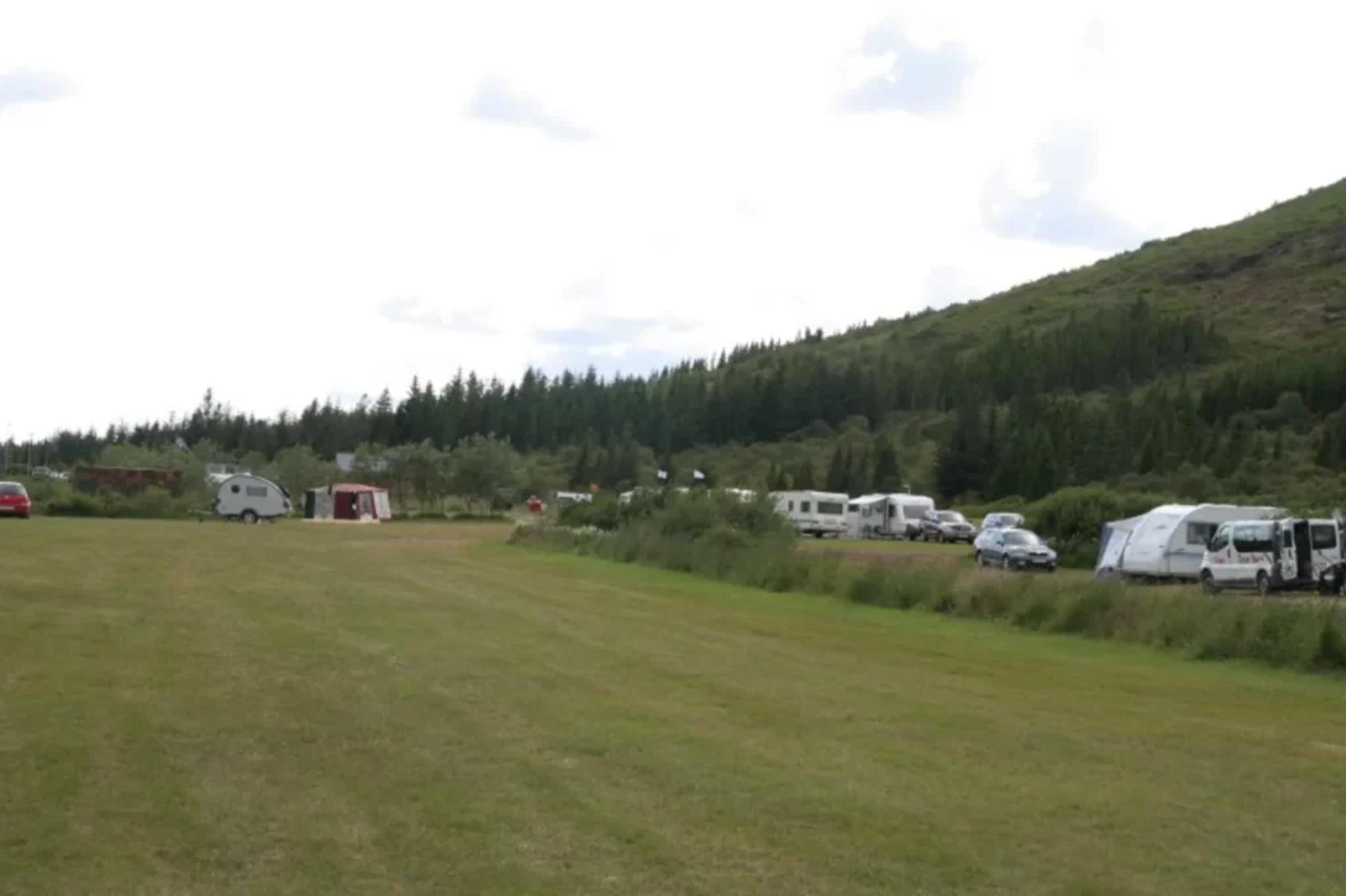 A grassy campsite with RVs, caravans, and tents, backed by a forest-covered hill.