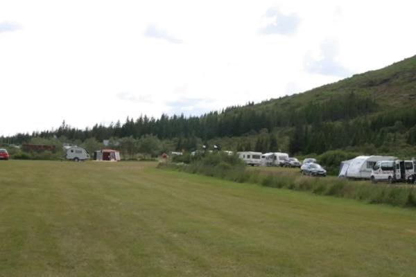 A grassy campsite with RVs, caravans, and tents, backed by a forest-covered hill.