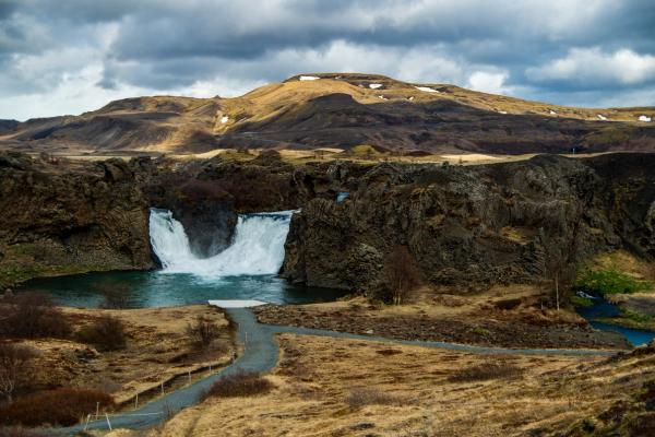 A wide waterfall plunges into a turquoise pool, surrounded by dark rocky cliffs and dry brown hills under a cloudy sky.