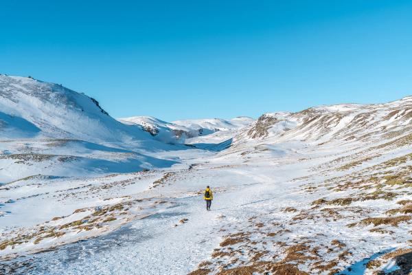 A person in a yellow jacket walks on a snowy path through a mountain valley under a clear blue sky.