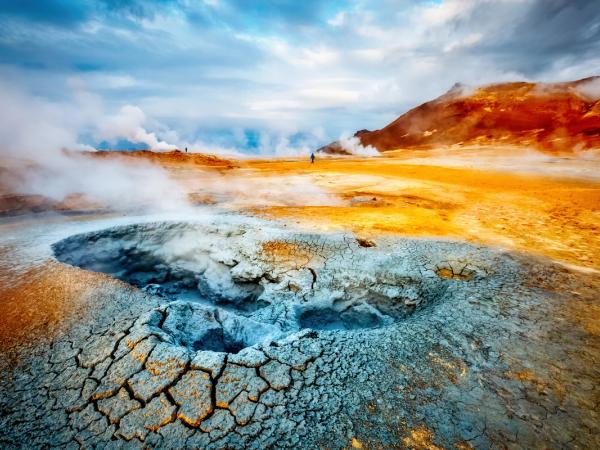 A steaming, cracked mud pot in a vibrant orange and grey geothermal landscape under a cloudy sky.