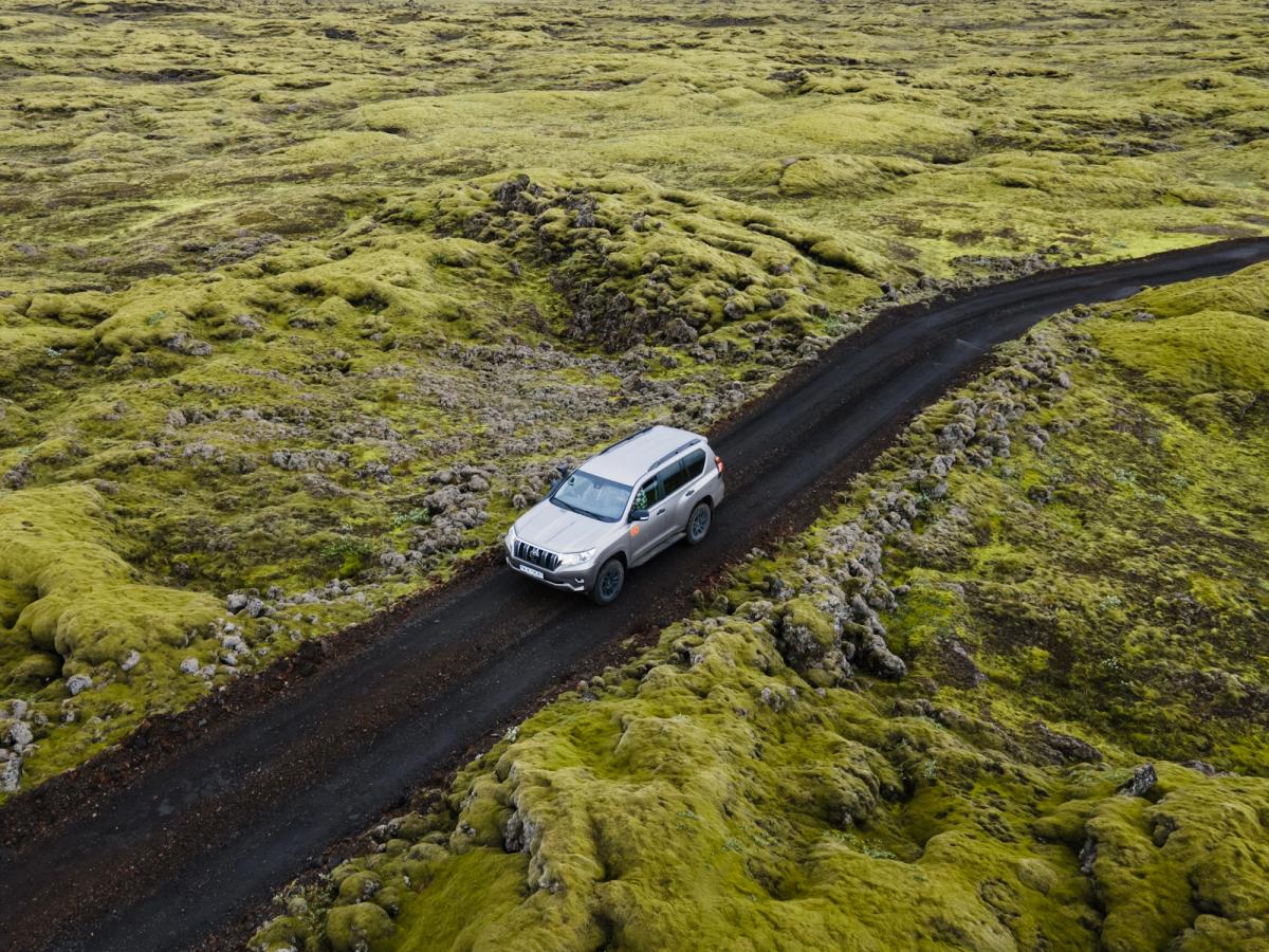 Loftmynd af fjórhjóladrifnum húsbíl á dökkum malarvegi um mosavaxið landslag, húsbílaferð um Ísland