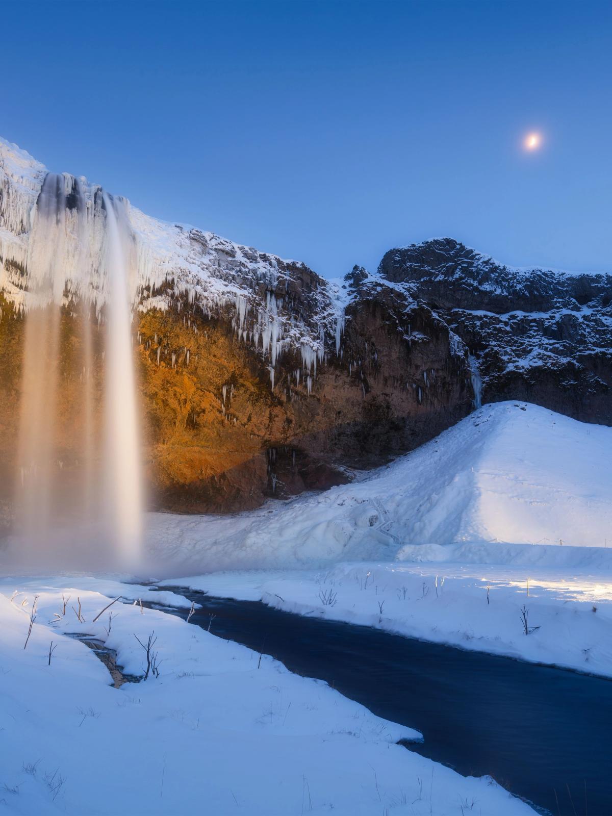 A waterfall cascades over icy, golden-lit cliffs into a snowy landscape under a blue twilight sky with a crescent moon.