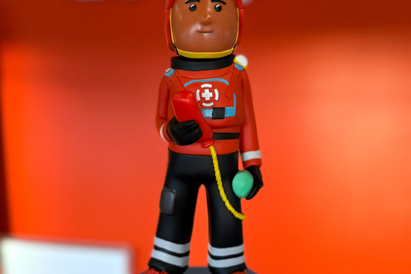A hand holds a figurine of a dark-skinned rescue worker in a red helmet and a red and black suit with a cross emblem, standing on a blue base with Icelandic text.