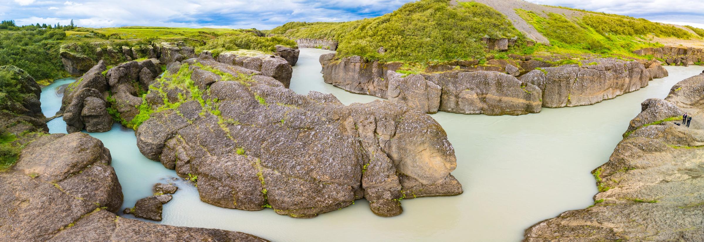 Una vista panorámica de un río con agua de color azul lechoso fluyendo a través de un cañón de grandes rocas grises con vegetación verde.