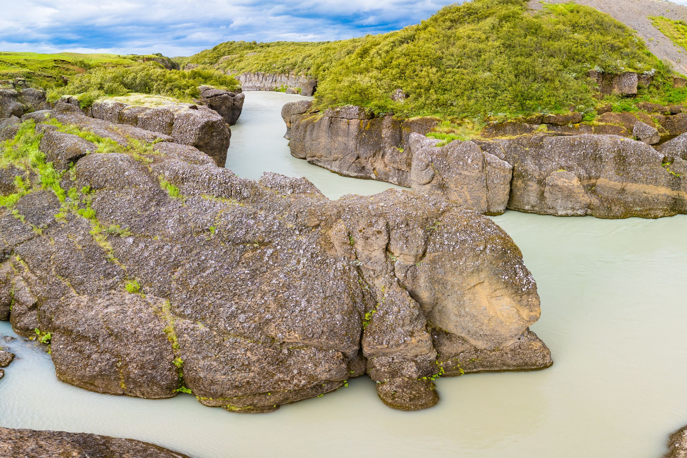 Una vista panorámica de un río con agua de color azul lechoso fluyendo a través de un cañón de grandes rocas grises con vegetación verde.