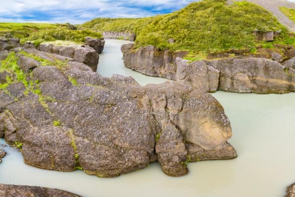 A panoramic view of a river with milky blue water flowing through a canyon of large grey rocks with green vegetation.