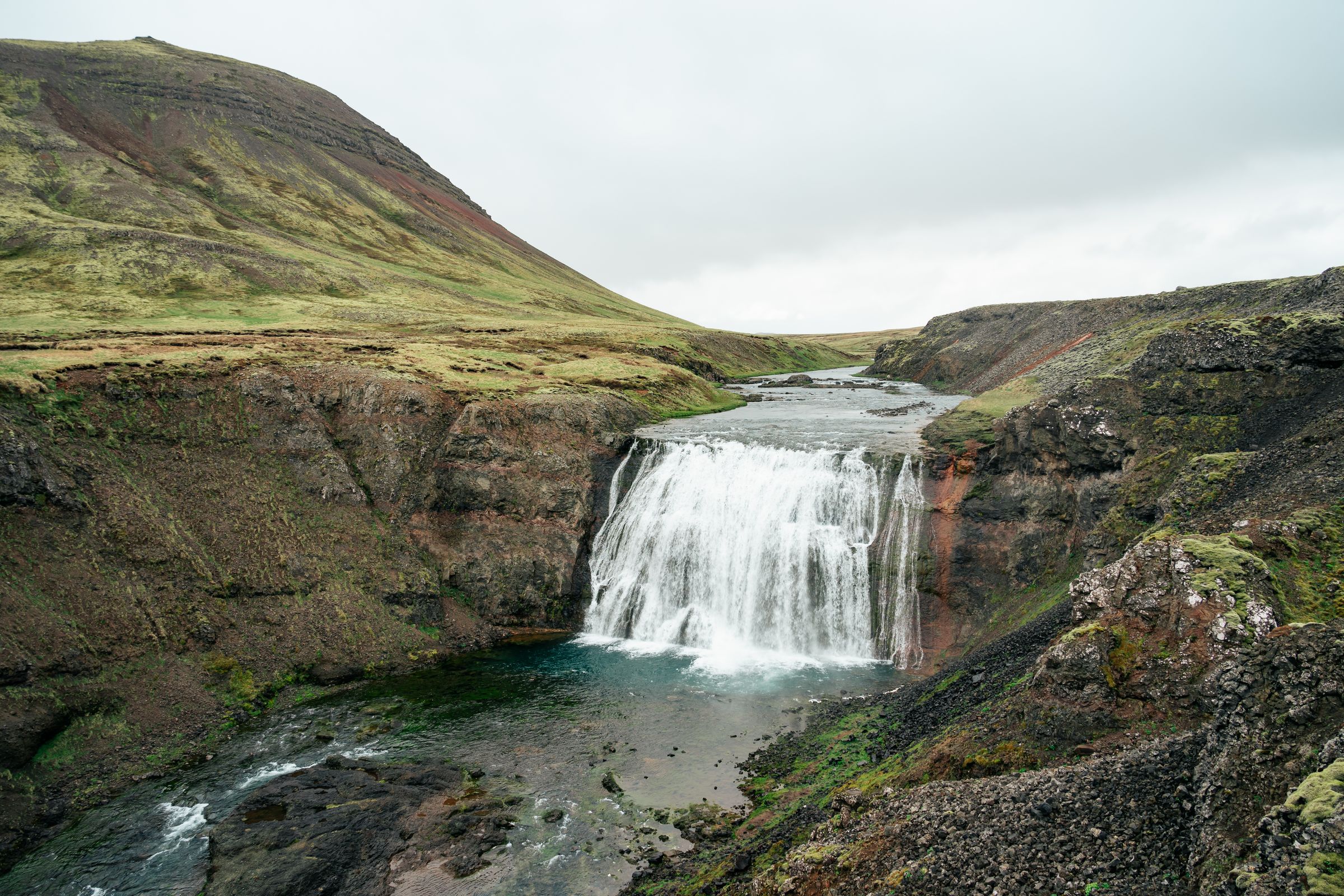 Waterfall in a rugged, green landscape under an overcast sky.