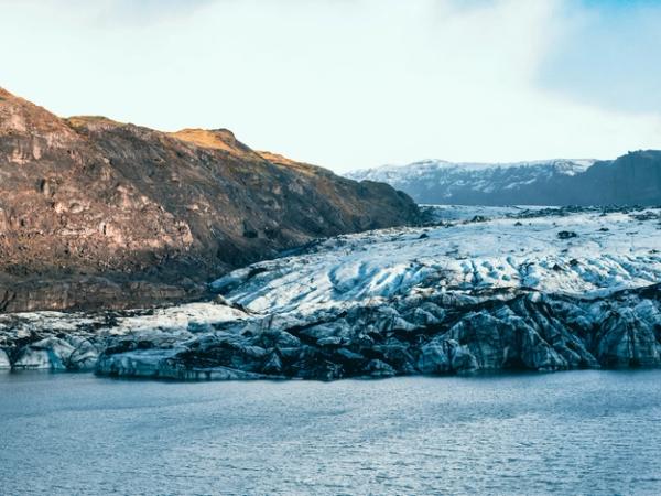Panoramic view of a large glacier extending into a lake, with brown mountains under a cloudy sky.