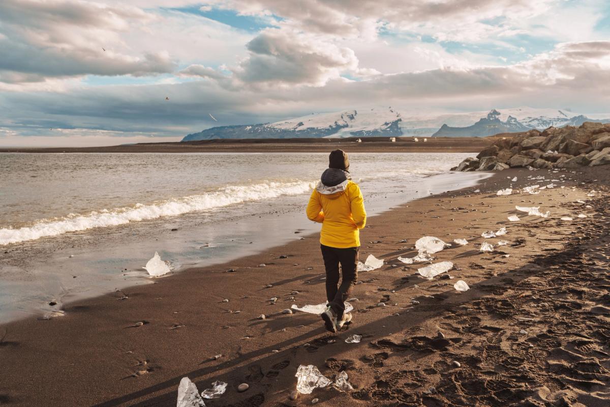 A person in a yellow jacket walks on a black sand beach scattered with ice chunks, with an ocean and snowy mountains in the background.