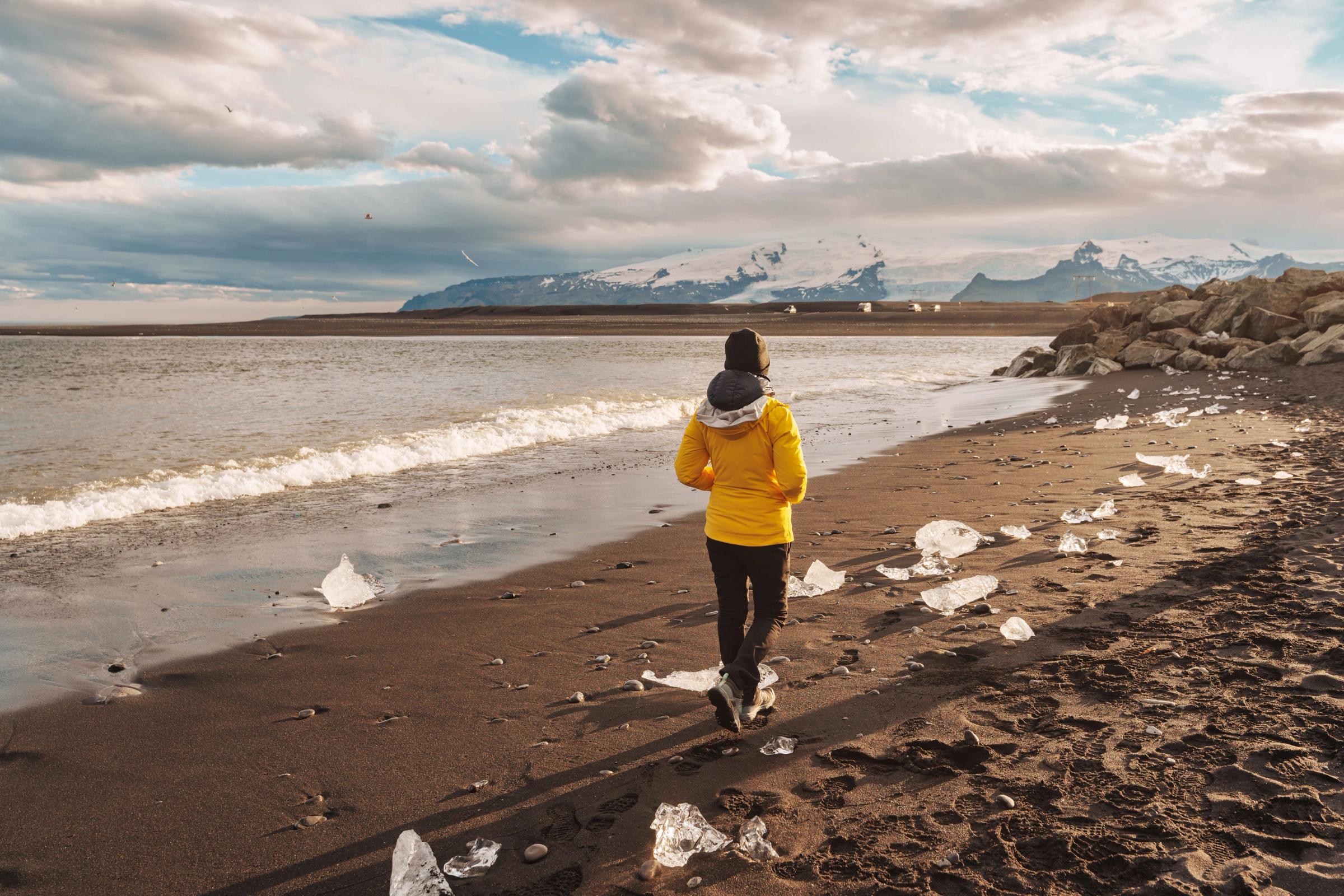Una persona con una chaqueta amarilla camina por una playa de arena negra salpicada de hielo, con el océano y montañas nevadas de fondo.