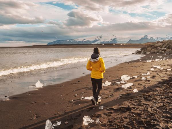 Una persona con una chaqueta amarilla camina por una playa de arena negra salpicada de hielo, con el océano y montañas nevadas de fondo.