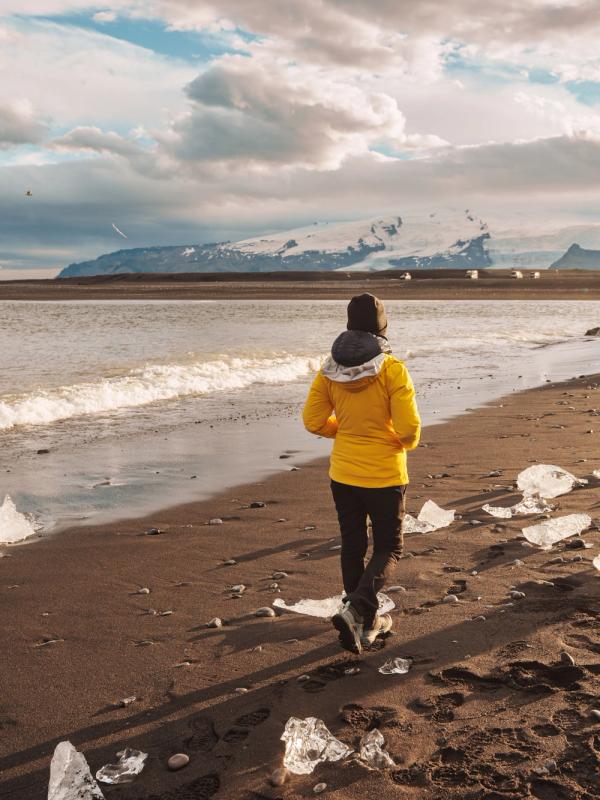 A person in a yellow jacket walks on a black sand beach scattered with ice chunks, with an ocean and snowy mountains in the background.