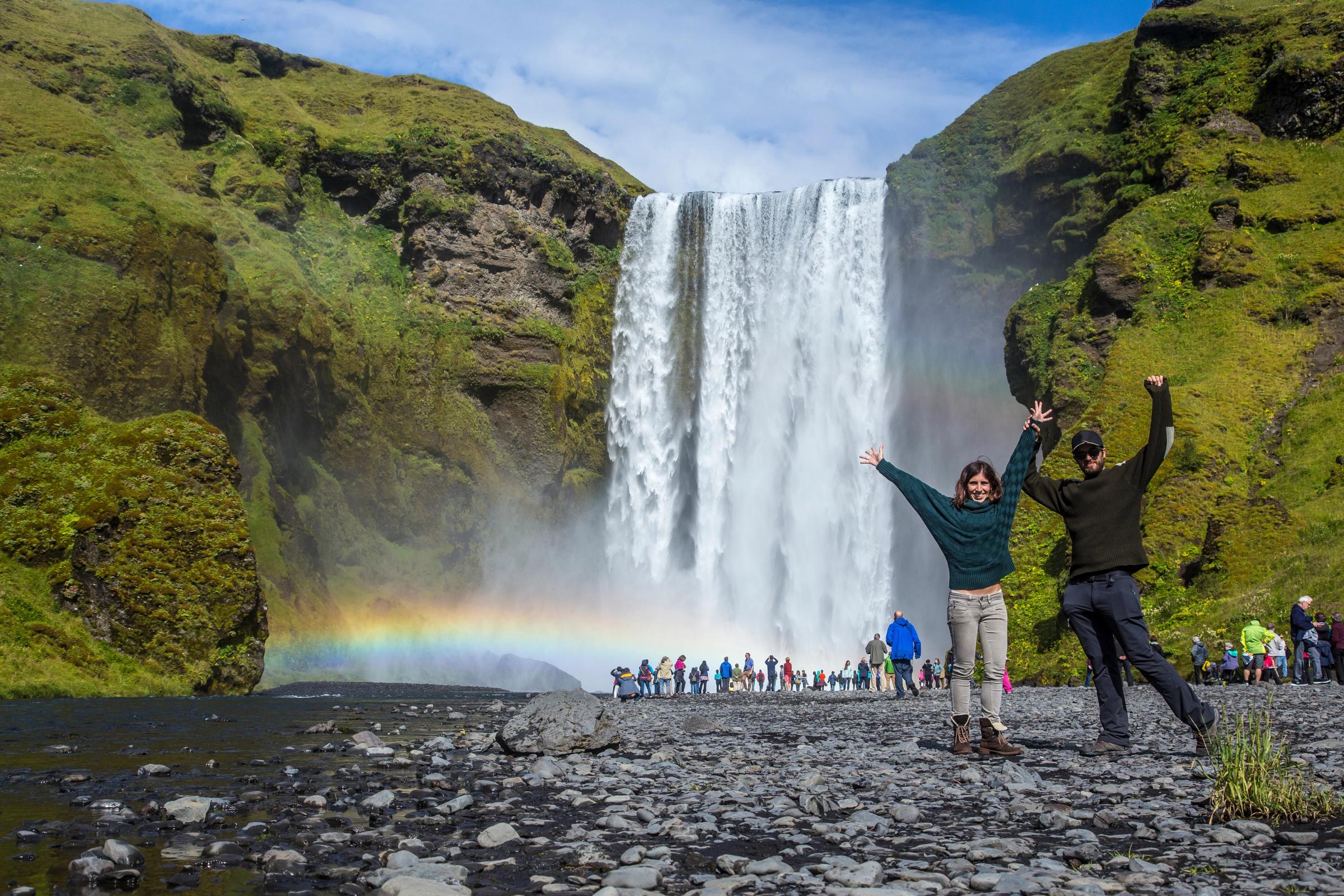 A massive waterfall cascades over green cliffs as two people cheer with a rainbow in the mist.