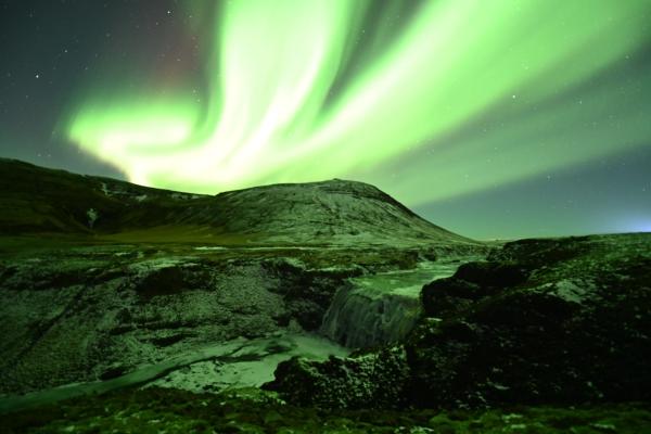 Green aurora borealis over snowy mountains with a river and waterfall.