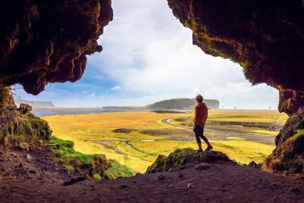 A person in an orange jacket stands at a cave entrance, looking out at a landscape of golden fields, a body of water, and distant cliffs under a bright sky.