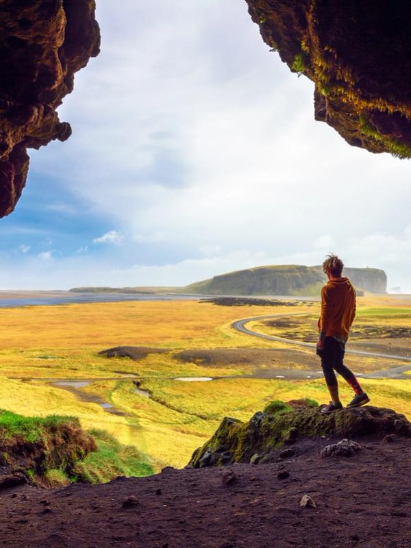 A person stands at a dark cave opening, looking out at a vast golden field and distant cliffs under a bright sky.