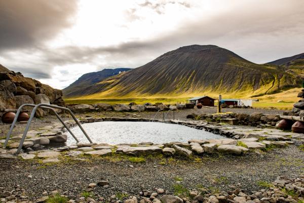 An outdoor hot spring pool with stone border and metal handrails, set against a backdrop of mountains and fields.