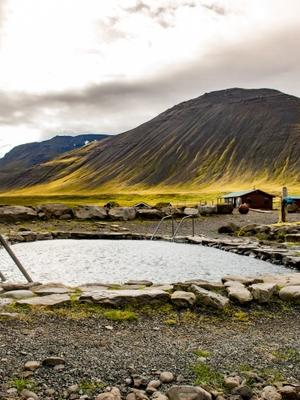 An outdoor hot spring pool with stone border and metal handrails, set against a backdrop of mountains and fields.