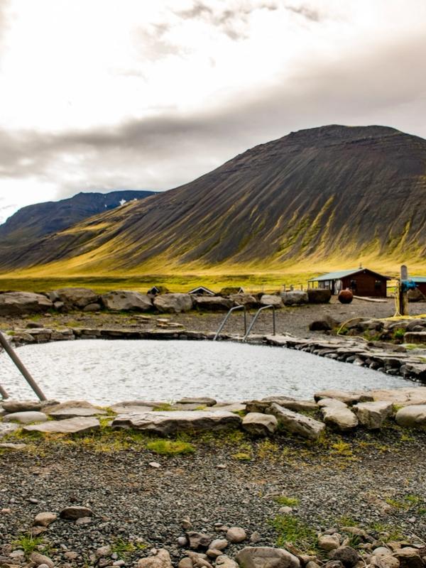 An outdoor hot spring pool with stone border and metal handrails, set against a backdrop of mountains and fields.