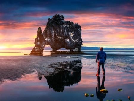 un homme se tient debout sur une plage en regardant l'Elephant Rock en Islande