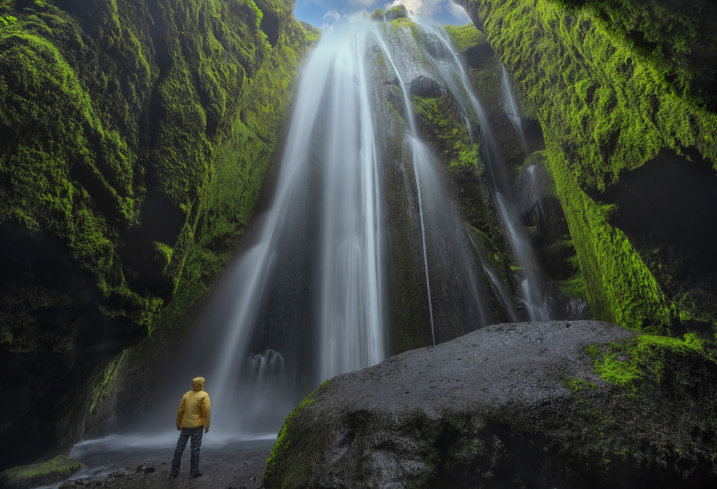 A person in a yellow jacket stands at the base of a tall, misty waterfall surrounded by green, moss-covered rock walls.