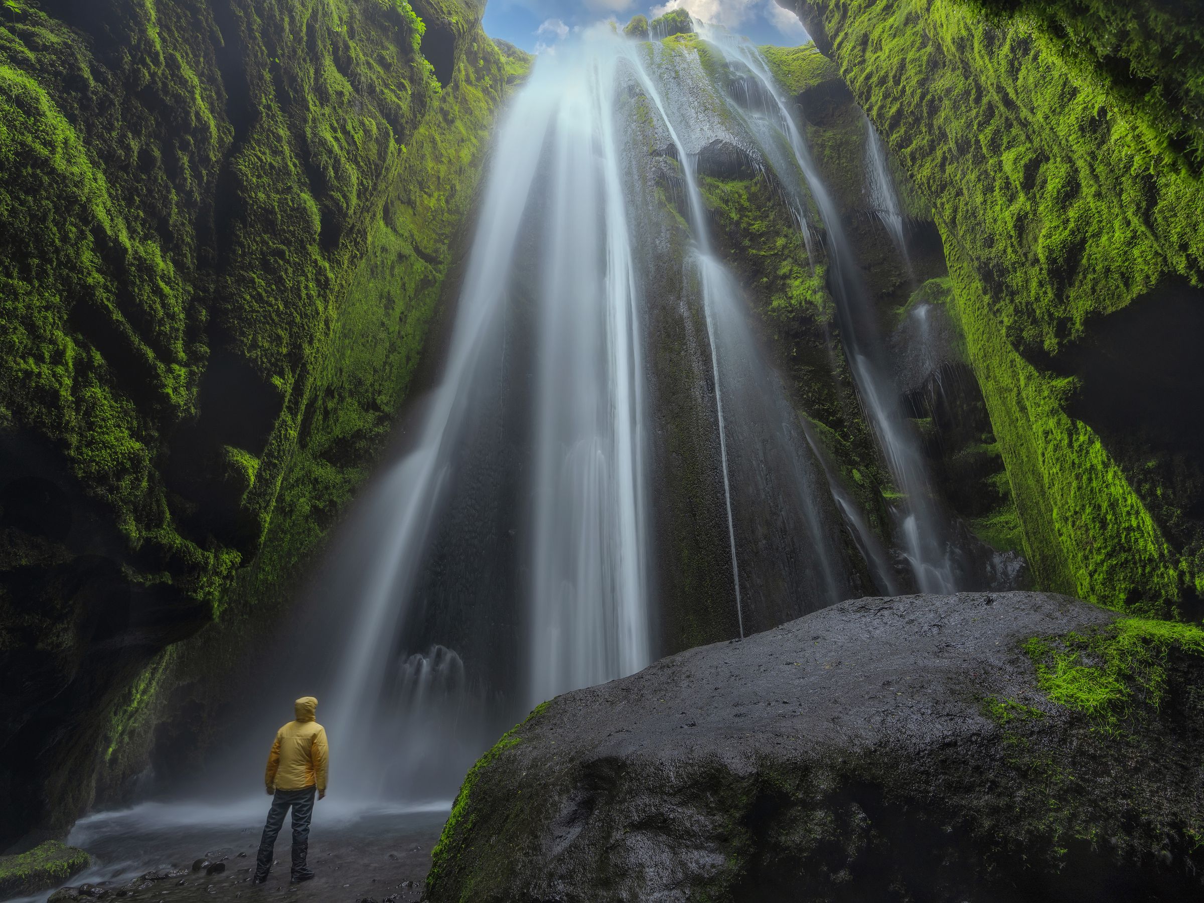 Gljúfrabúi A person in a yellow jacket stands at the base of a tall, misty waterfall surrounded by green, moss-covered rock walls.