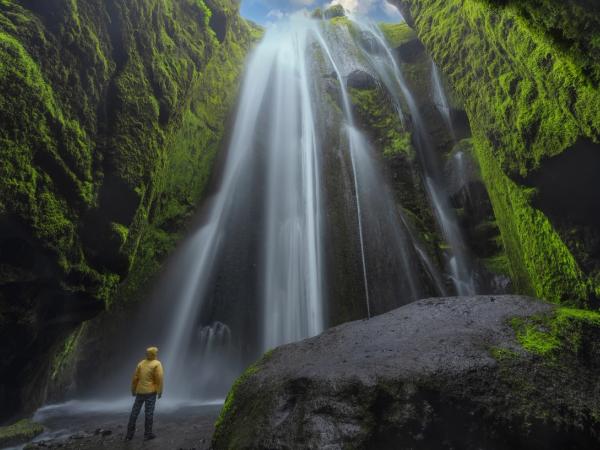 A person in a yellow jacket stands before a tall, silky waterfall within a moss-covered canyon.