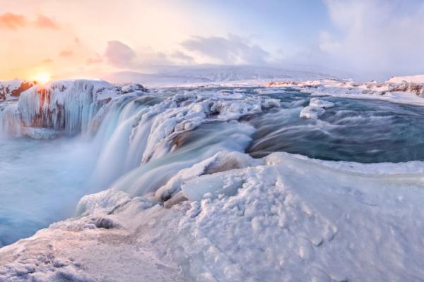 Panoramic view of an icy waterfall surrounded by snow, with a warm sunset glow in the sky.
