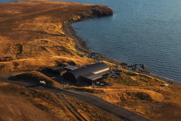 Aerial view of a dark, modern building with a curved roof on a golden-brown, arid coastline next to blue water.