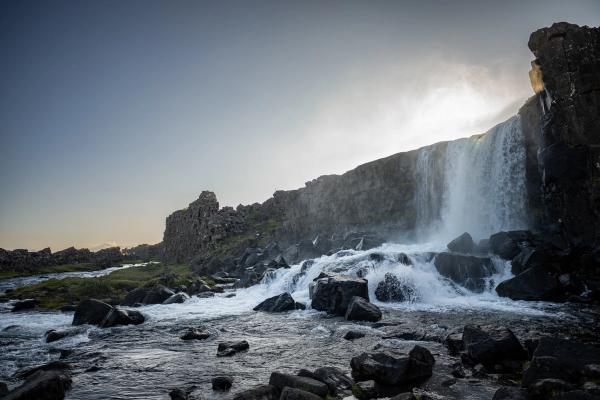 A powerful waterfall cascades over dark rocky cliffs into a rocky river, with bright sun behind the falls.