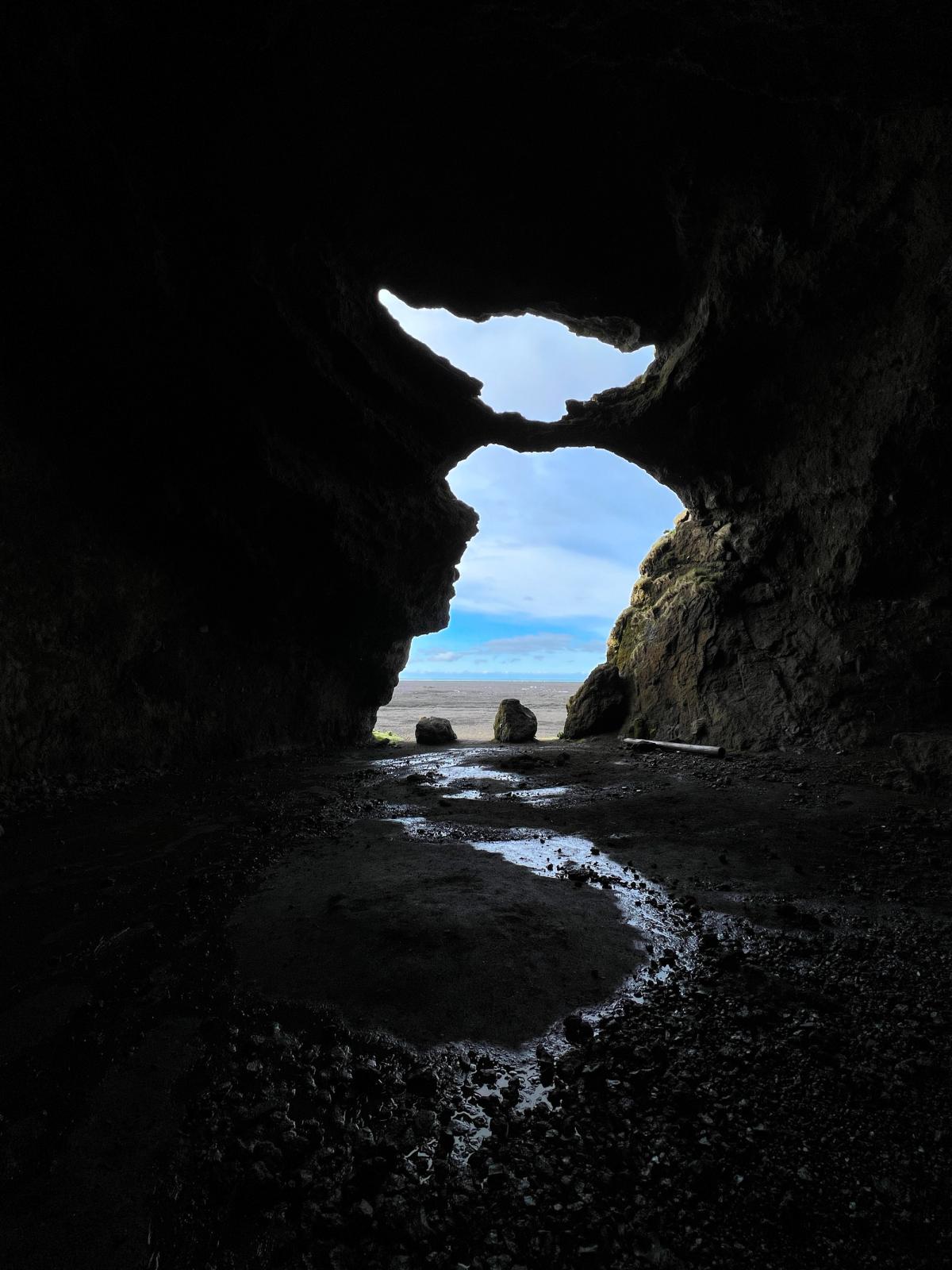 Looking out from a dark cave through natural rock arches to a bright blue sky and distant ocean over wet ground.