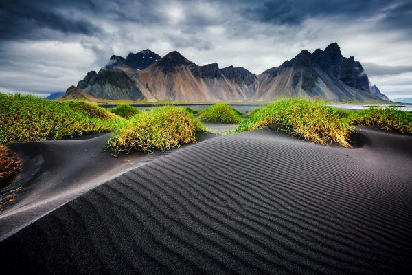 a desert landscape with mountains in the background and a black sand dune in the foreground . vestrarhorn by höfn in iceland.