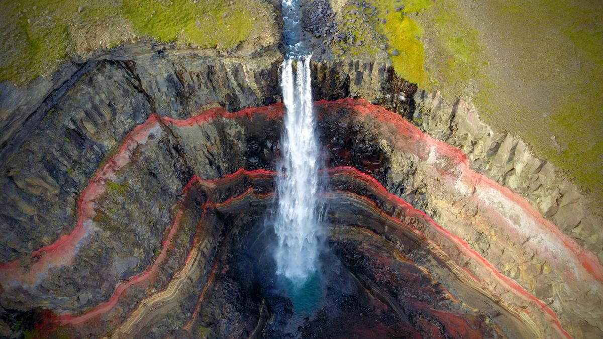 A waterfall cascades down a cliff face revealing prominent red and multicolored geological layers.